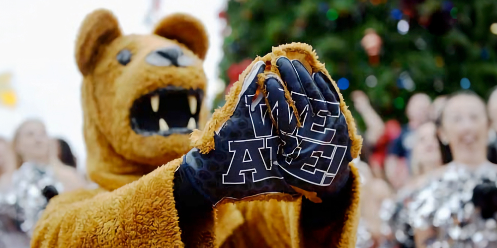 Nittany Lion mascot holds out hands that have "WE ARE" written on them.
