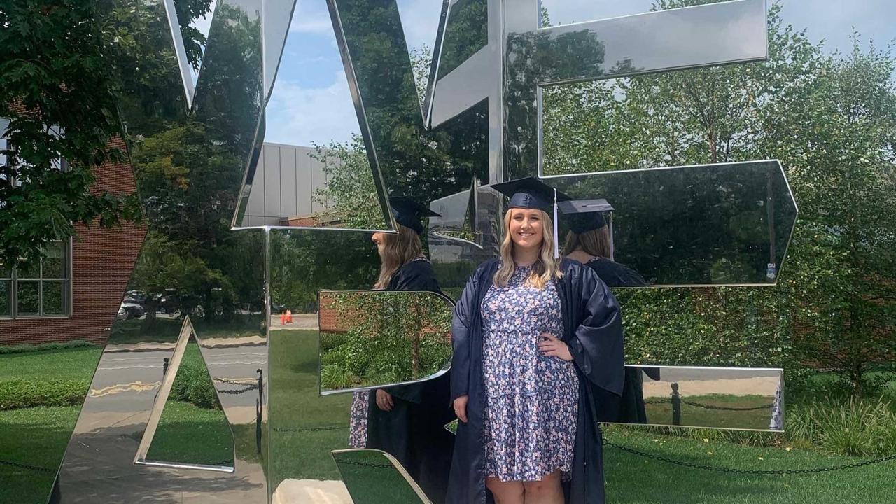 Abigail Bauer stands in front of the We Are sculpture on the Penn State University Park campus, smiling and wearing her graduation cap